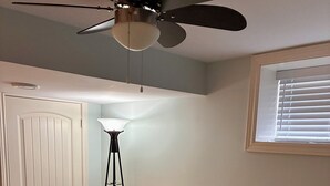 Ett sovrum med en säng, en golvlampa, en takfläkt, ett fönster med persienner och en dörr.