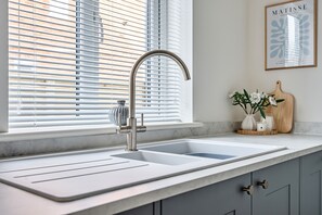 A kitchen with a modern faucet, a window with blinds, a framed artwork on the wall, and a wooden cutting board with a vase of flowers on the counter. - Beautifully renovated cottage in a fantastic location with free parking (Stratford-upon-Avon)