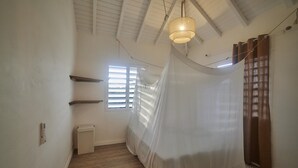 Um quarto com piso de madeira, ventilador de teto, janela com persianas e uma cama com dossel branco, além de uma prateleira fixada na parede.
