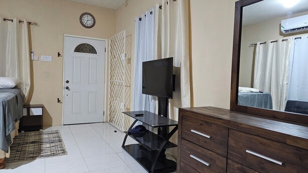 A hotel room with a bed, a TV, a dresser, a mirror, a door, and a clock on the wall. - Tandel Ocho Rios Apartment #2 (Ocho Rios)