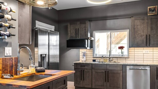 Cocina moderna con armarios oscuros, electrodomésticos de acero inoxidable, encimera de madera y ventana con vistas.