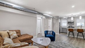 Una sala de estar moderna con un sofá de cuero marrón, una mesa de centro blanca y un sillón azul. Se ve una cocina con gabinetes blancos y un microondas al fondo.