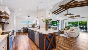 A modern kitchen with a large island, wooden flooring, pendant lighting, and a view of the living area with a sofa and a TV. - Bahama Mama | Tropical Retreat on Siesta Key (Siesta Key)