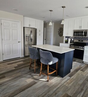 A modern kitchen with white cabinets, a stainless steel refrigerator, a black stove, and two bar stools.