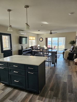 A modern kitchen with a central island, dining area with chairs, a ceiling fan, and a large window with a view of the outdoors.