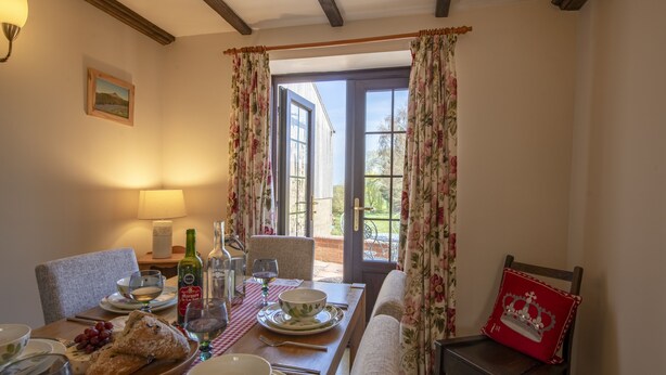 A dining area with a table set for two, a bench, a lamp, and a view through an open door. - Lyke Wake Cottage, Swainby, Yorkshire (Northallerton)