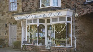 A brick building with a shop named 'Swanby Village Store' featuring large windows, a door, and outdoor seating. - Lyke Wake Cottage, Swainby, Yorkshire (Northallerton)