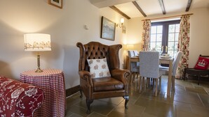 A room with a leather armchair, a round table with a checkered tablecloth, a dining table with chairs, and a window with curtains. - Lyke Wake Cottage, Swainby, Yorkshire (Northallerton)