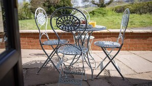 A patio with metal chairs and a table, a brick wall, and a view of a grassy landscape. - Lyke Wake Cottage, Swainby, Yorkshire (Northallerton)
