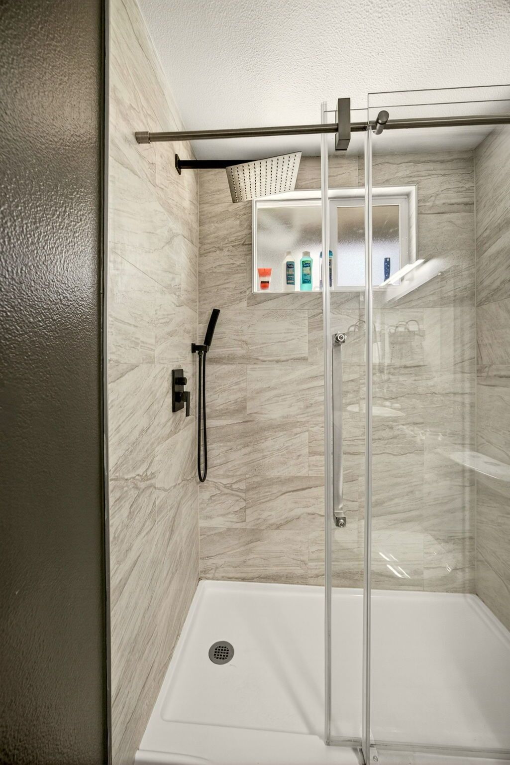Una ducha moderna con puerta de cristal, cabezal de ducha manual y repisa con artículos de aseo.