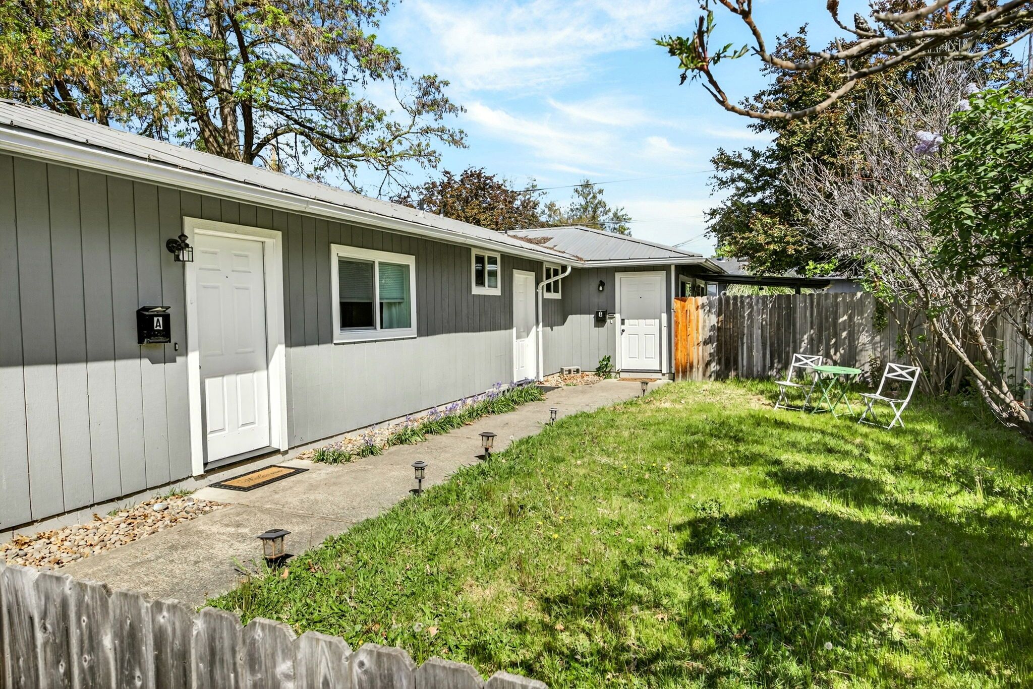 A single-story house with a grey exterior, a front door, and a small front yard with a wooden fence and a bench.