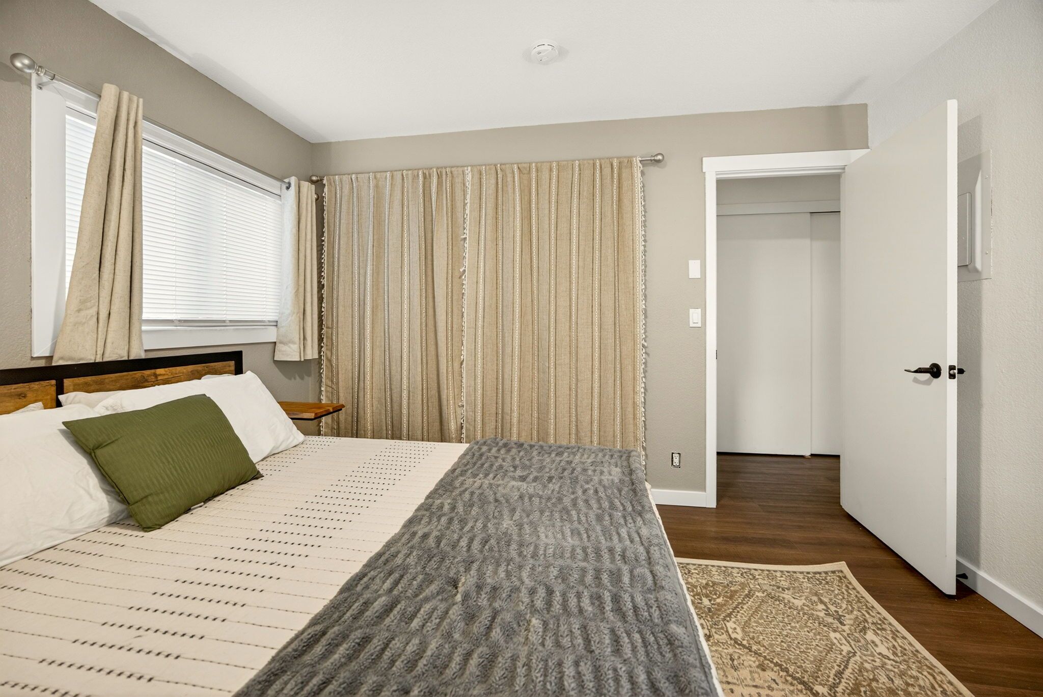 A bedroom with a bed, a nightstand, a window with curtains, a door leading to another room, and a patterned rug.