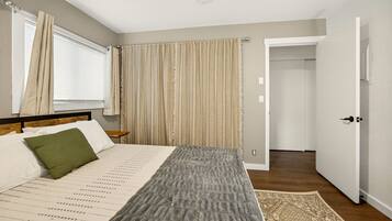 A bedroom with a bed, a nightstand, a window with curtains, a door leading to another room, and a patterned rug.