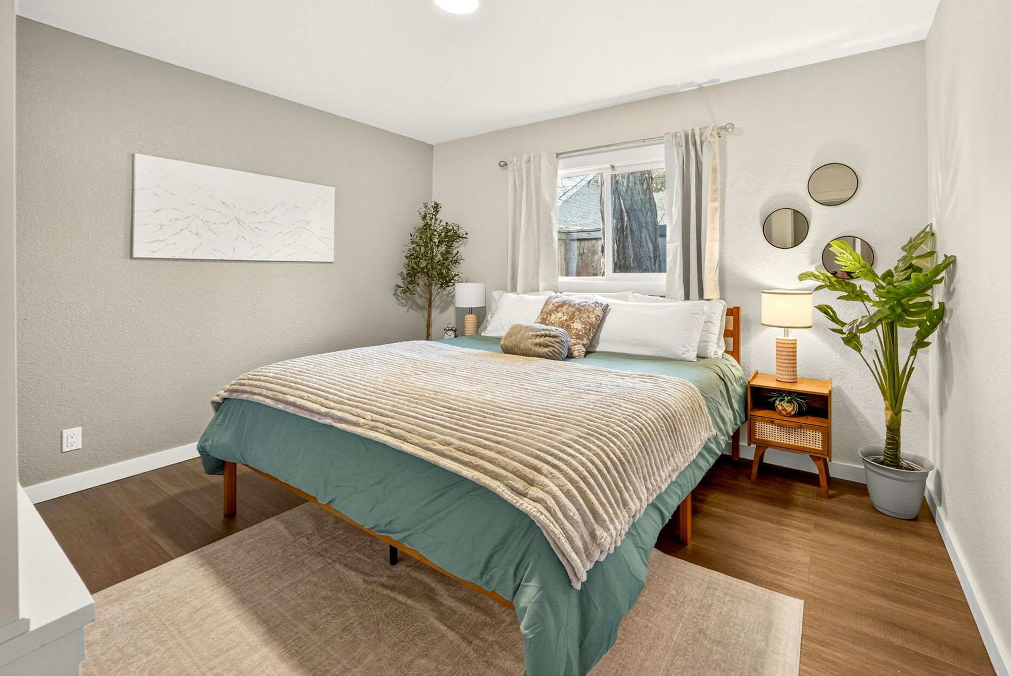 A bedroom with a bed, bedside tables, a window with curtains, a potted plant, and a wall-mounted artwork.