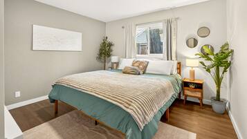A bedroom with a bed, bedside tables, a window with curtains, a potted plant, and a wall-mounted artwork.