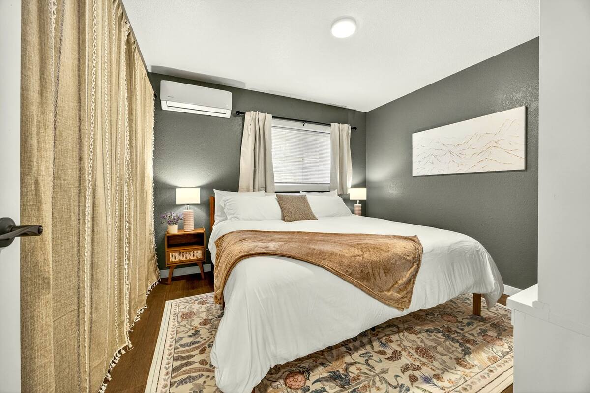 Un dormitorio con cama, mesitas de noche, ventana con cortinas, equipo de aire acondicionado y un cuadro colgado en la pared.