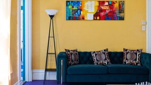 A teal sofa with patterned cushions, a floor lamp, a chessboard, and a colorful abstract painting on the wall. - Retro 1BR Flat in the Heart of East Liberty | Queen Bed | Longer Stays Welcome (Pittsburgh)