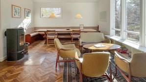 Une pièce dotée d’un parquet en bois à motifs en chevrons, d’une cheminée, d’une table à manger avec des chaises, d’une fenêtre donnant sur des arbres et d’une lampe.