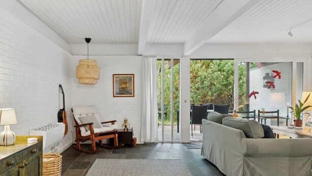 A modern living room with a white brick wall, a wooden rocking chair, a sofa, a dining table with chairs, and a large glass door leading to an outdoor area.