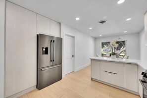 A modern kitchen with a stainless steel refrigerator, white cabinetry, and a window with a view of trees. - waterfront corner lot (Pompano Beach)