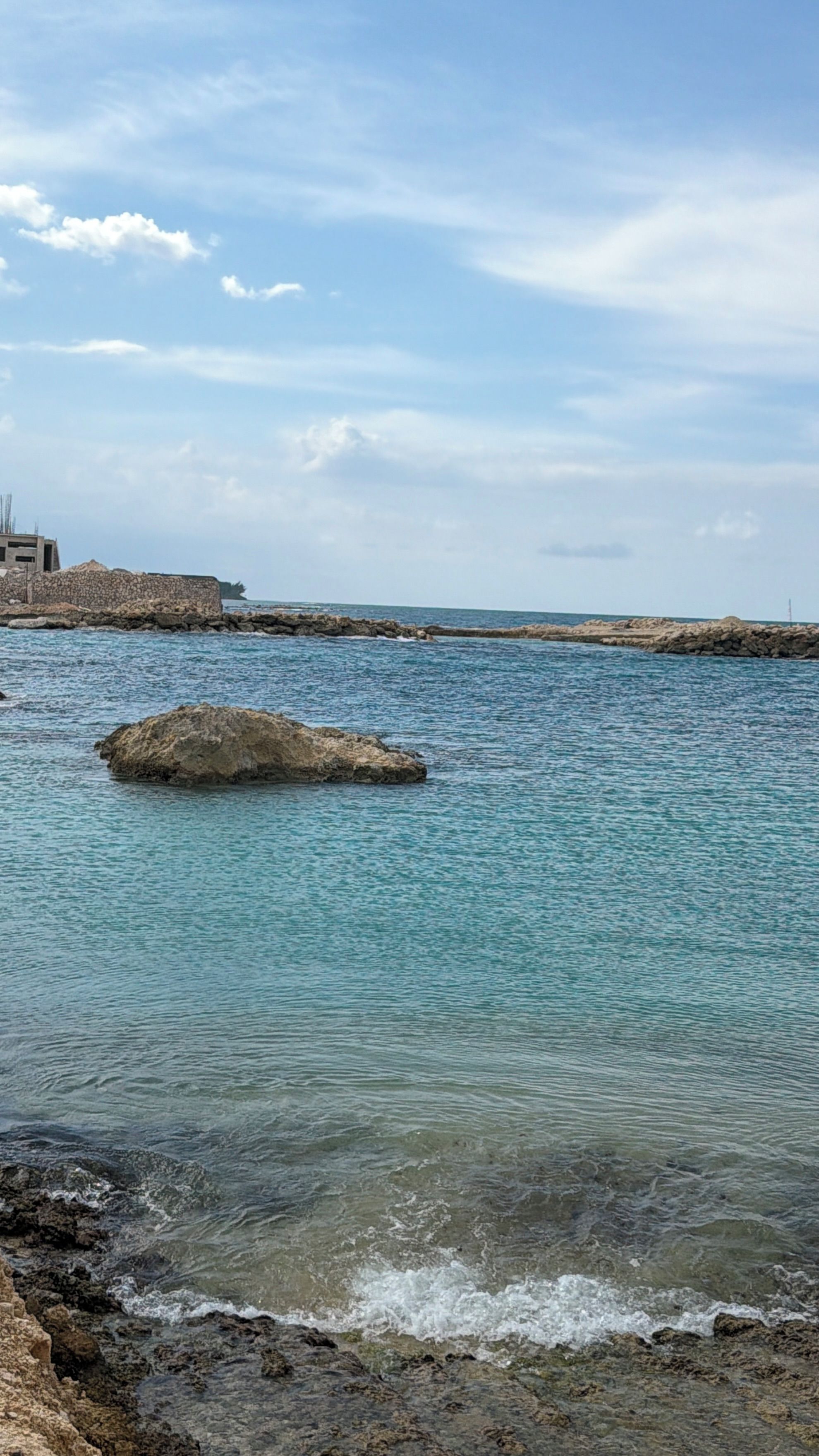 A coastal scene with clear water, a large rock in the center, and rocky shorelines.
