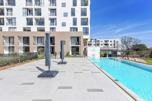A modern building with a swimming pool, outdoor seating, and a clear sky. - One Rosebank | Calm. Luxe. Living. (Johannesburg)