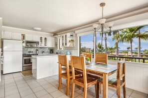 A kitchen with white cabinets, a dining area with wooden chairs, and a view of palm trees through large windows.