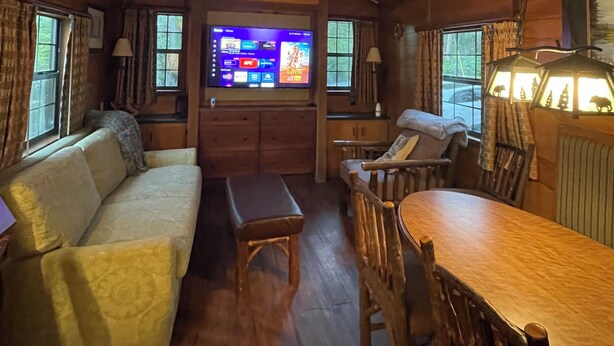 Ein gemütliches Zimmer mit Holzbalkendecke, einem Fernseher, einem Sofa, einem Esstisch und einem Stuhl.