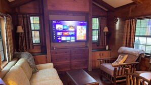 Ein gemütliches Zimmer mit einem großen Flachbildfernseher, der an einer Holzwand angebracht ist, einer Couch, einer Bank und zwei Holzstühlen.