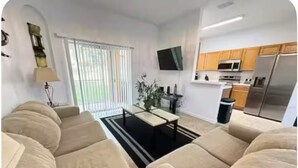 A living room with beige sofas, a coffee table, a TV mounted on the wall, and a kitchen area with wooden cabinets and appliances. - 324 Palermo Villa 30 min to Disney and Universal (Davenport)