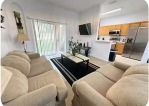 Ein Wohnzimmer mit beigefarbenen Sofas, einem Couchtisch, einem an der Wand befestigten Fernseher und einer Küchenzeile mit Holzschränken und Geräten.