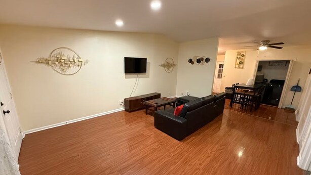 A spacious living room with a wooden floor, a black sofa, a television mounted on the wall, and a ceiling fan.