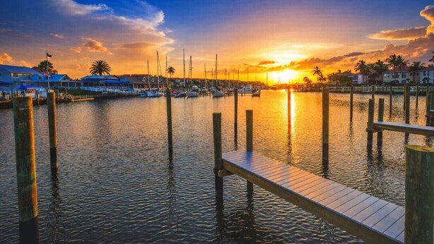 A marina at sunset with boats docked, wooden piers, and buildings in the background.