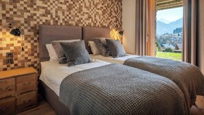 A double bed with a checkered wall, bedside lamps, a wooden nightstand, and a view of a village through a window.