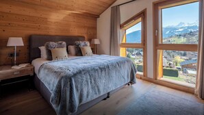A bedroom with a wooden ceiling, a large bed with a grey blanket, two bedside lamps, and a window with a mountain view.