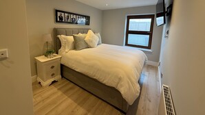 Une chambre à coucher comprenant un lit, une table de chevet, une lampe, des photos encadrées, une fenêtre et un téléviseur fixé au mur.