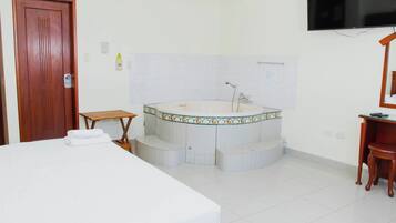Habitación de hotel con cama, una silla de madera, una mesita, bañera y televisión.