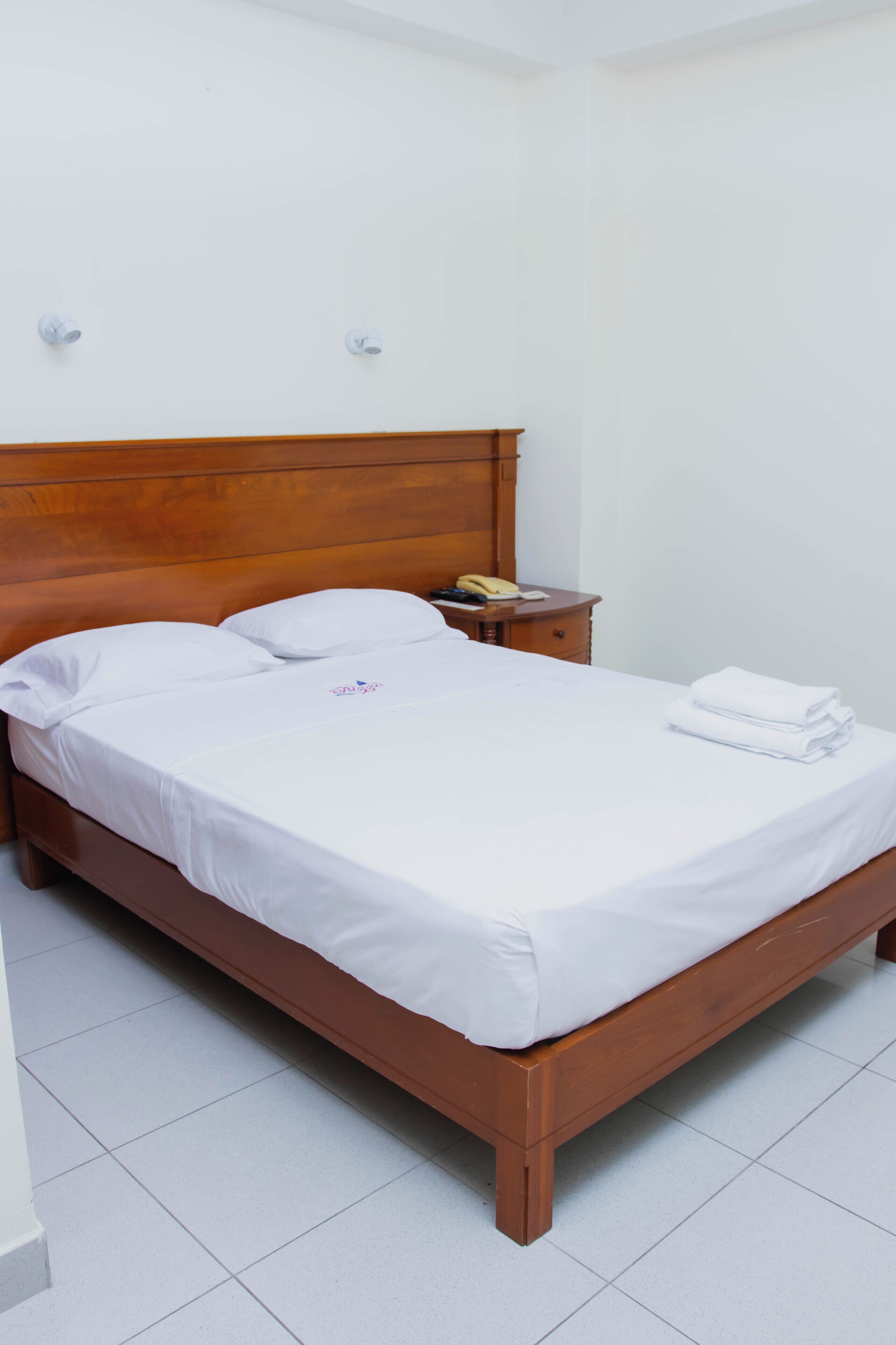 Una cama bien hecha con ropa de cama blanca, cabecera de madera, mesita de noche con lámpara y aplique de pared.