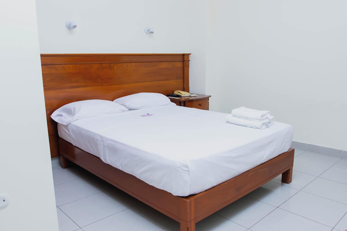 A neatly made bed with white linens, a wooden headboard, a bedside table with a lamp, and a wall-mounted light fixture.