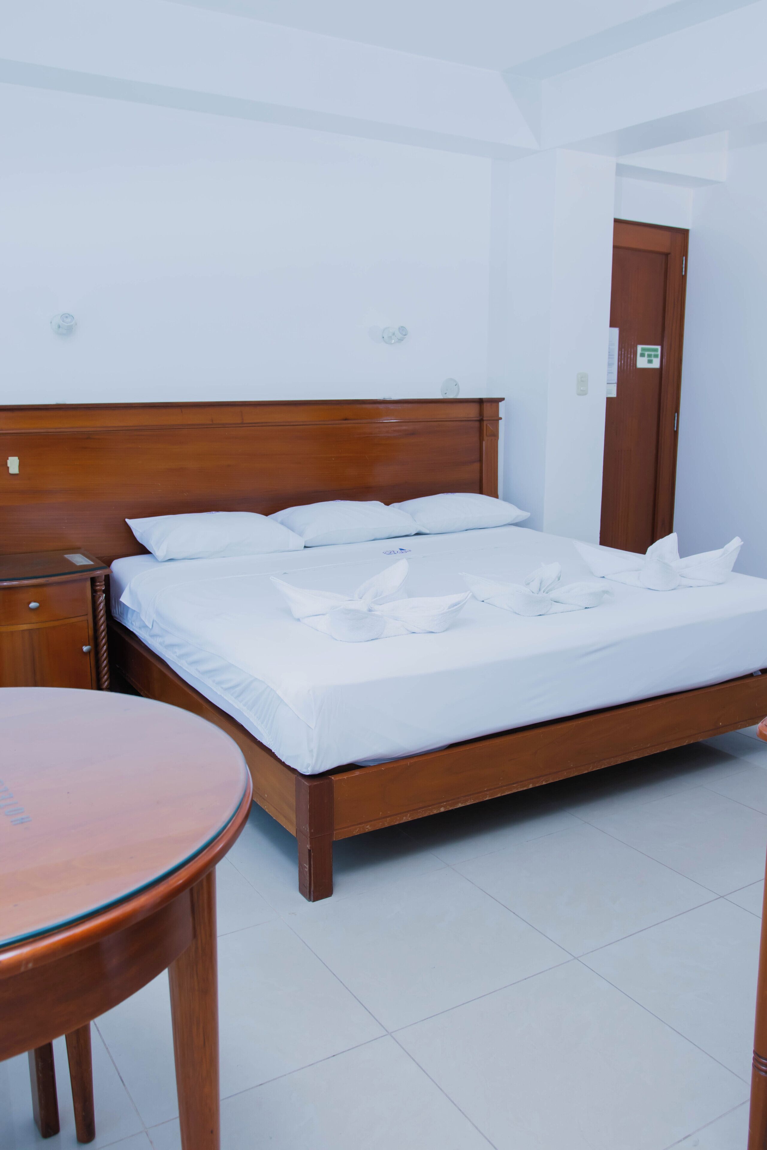 A neatly made bed with white linens and a wooden headboard, a wooden nightstand with a drawer, and a round wooden table.