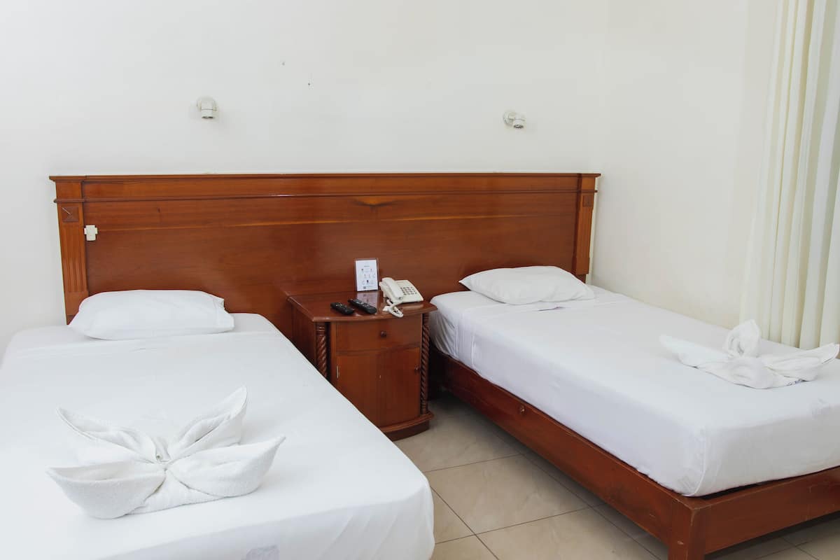 A hotel room with two single beds, a wooden headboard, a bedside table with a phone and remote controls, and a white towel on the beds.