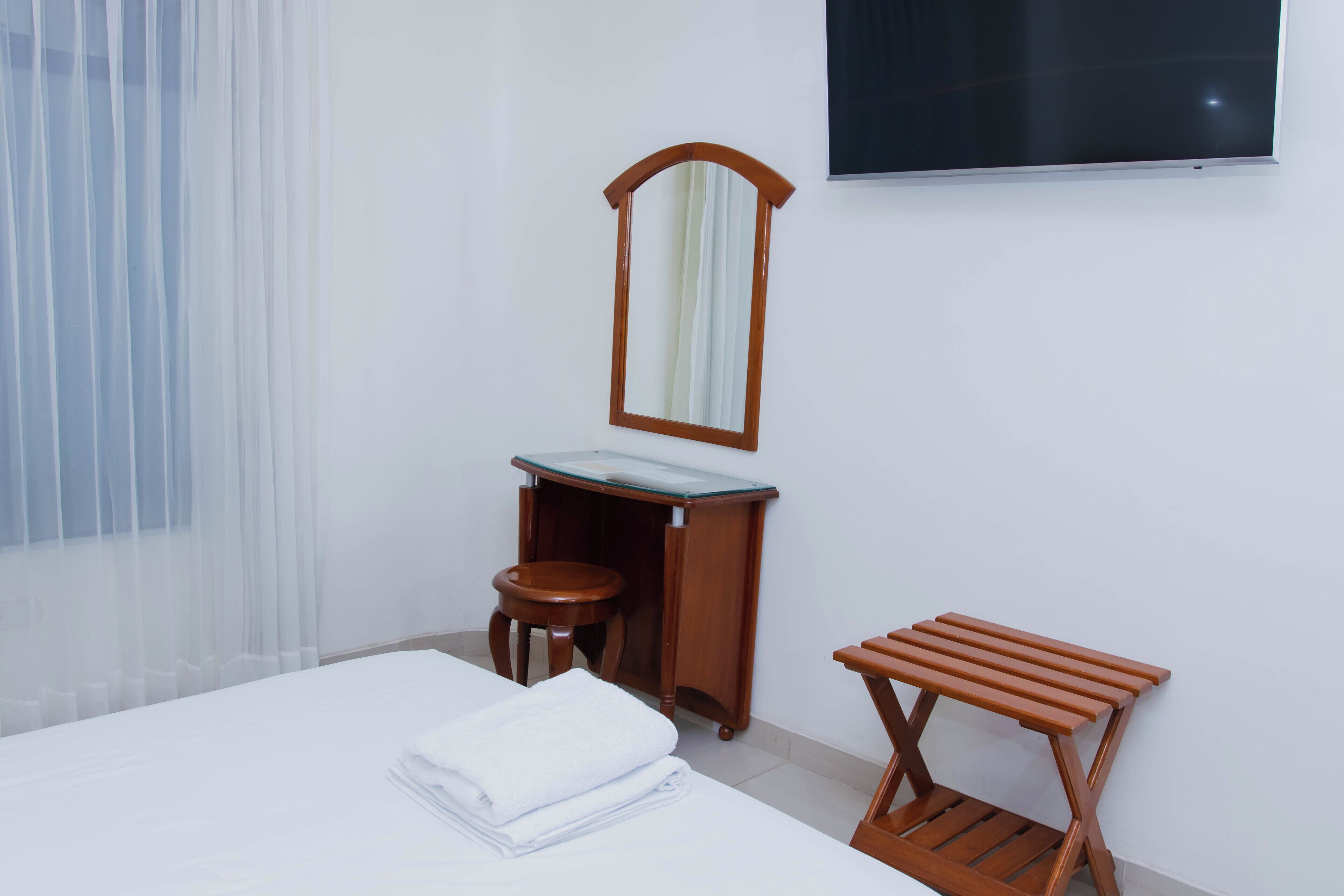 A hotel room with a bed, a wooden stool, a small table, a mirror, and a TV on the wall.