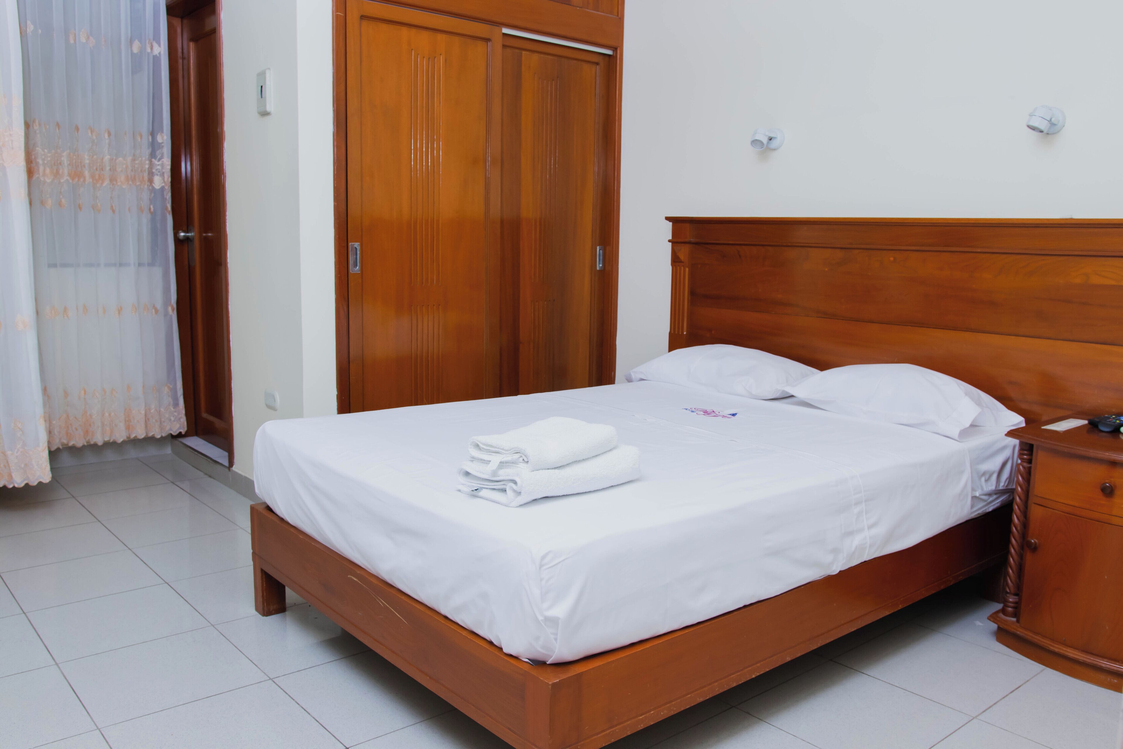 Un lit bien fait, avec du linge de lit blanc et une serviette pliée, une tête de lit en bois, une table de chevet en bois et une armoire en bois dotée d’une porte coulissante.