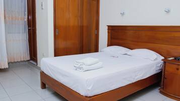 Un lit bien fait, avec du linge de lit blanc et une serviette pliée, une tête de lit en bois, une table de chevet en bois et une armoire en bois dotée d’une porte coulissante.
