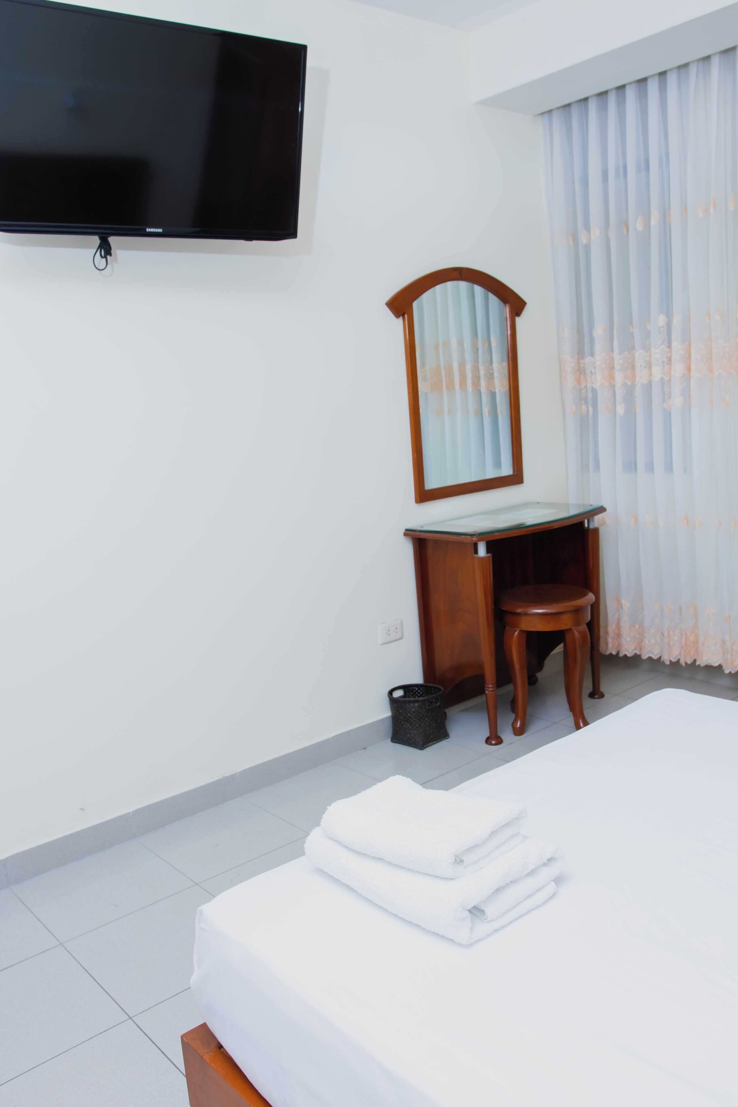Habitación de hotel con cama, televisor montado en la pared, escritorio de madera con espejo y ventana con cortinas traslúcidas.