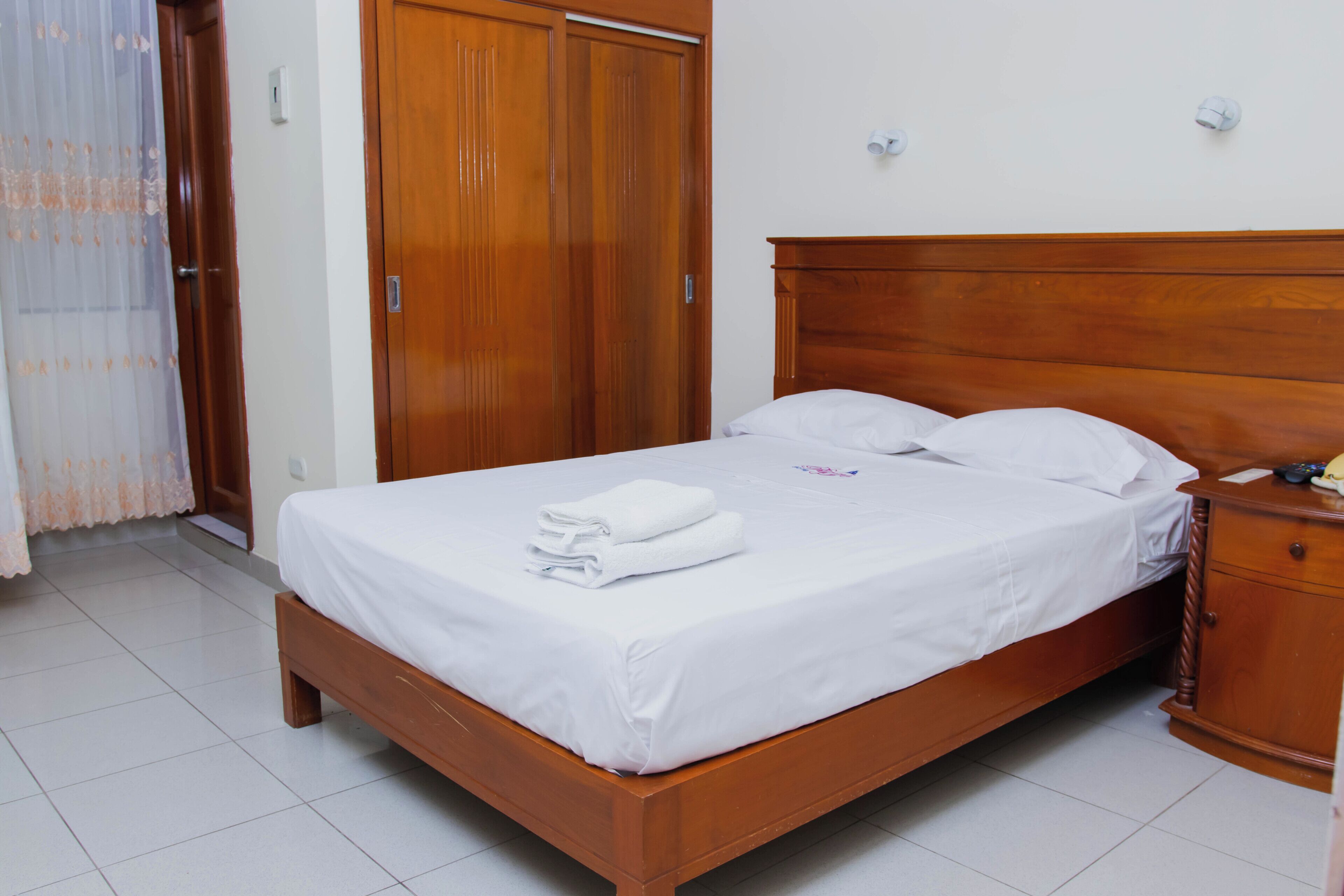 Una cama bien hecha con sábanas blancas, cabecera de madera y mesita de noche con una lámpara.
