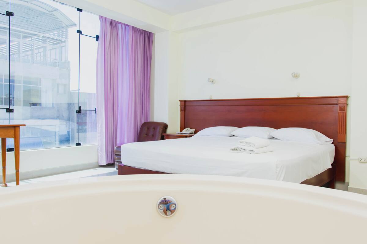 A hotel room with a large bed, a wooden headboard, a bedside table, a chair, and a bathtub.
