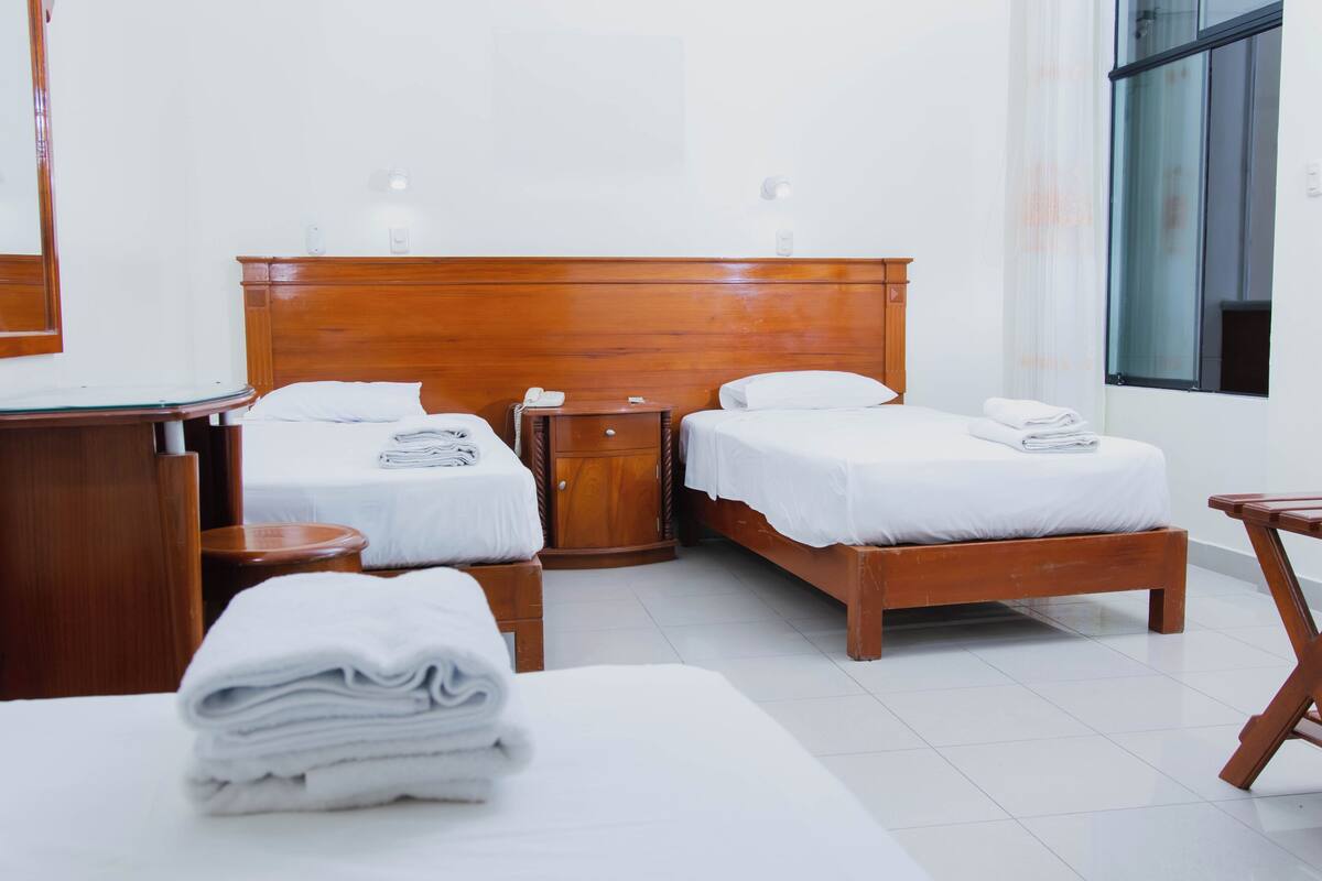 A hotel room with two beds, a wooden headboard, a bedside table with a lamp, a small wooden cabinet, and a window with a view.