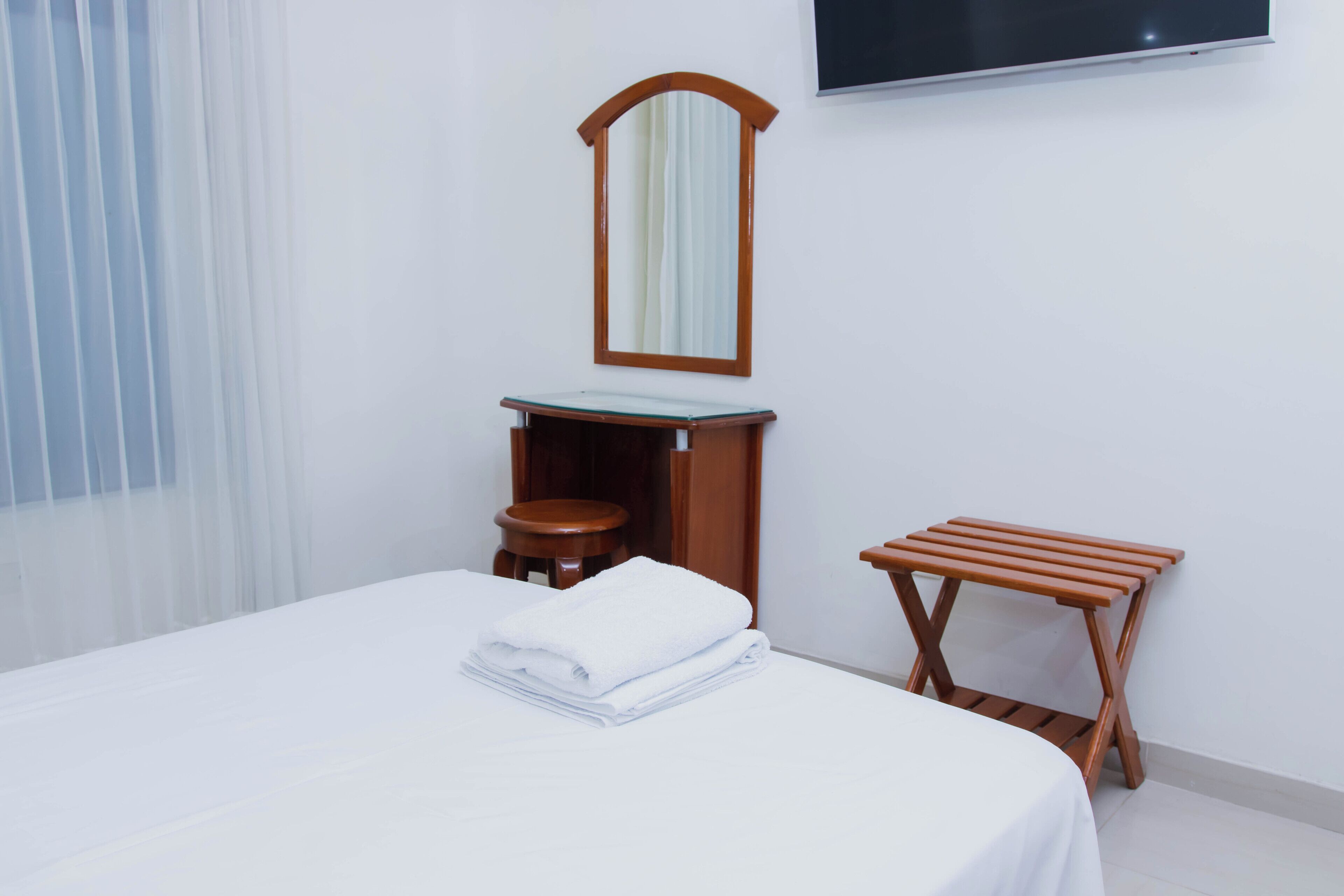 Ein Hotelzimmer mit einem Bett, einem Holzschemel, einem Nachttisch aus Holz, einem Spiegel und einem an der Wand befestigten Fernseher.