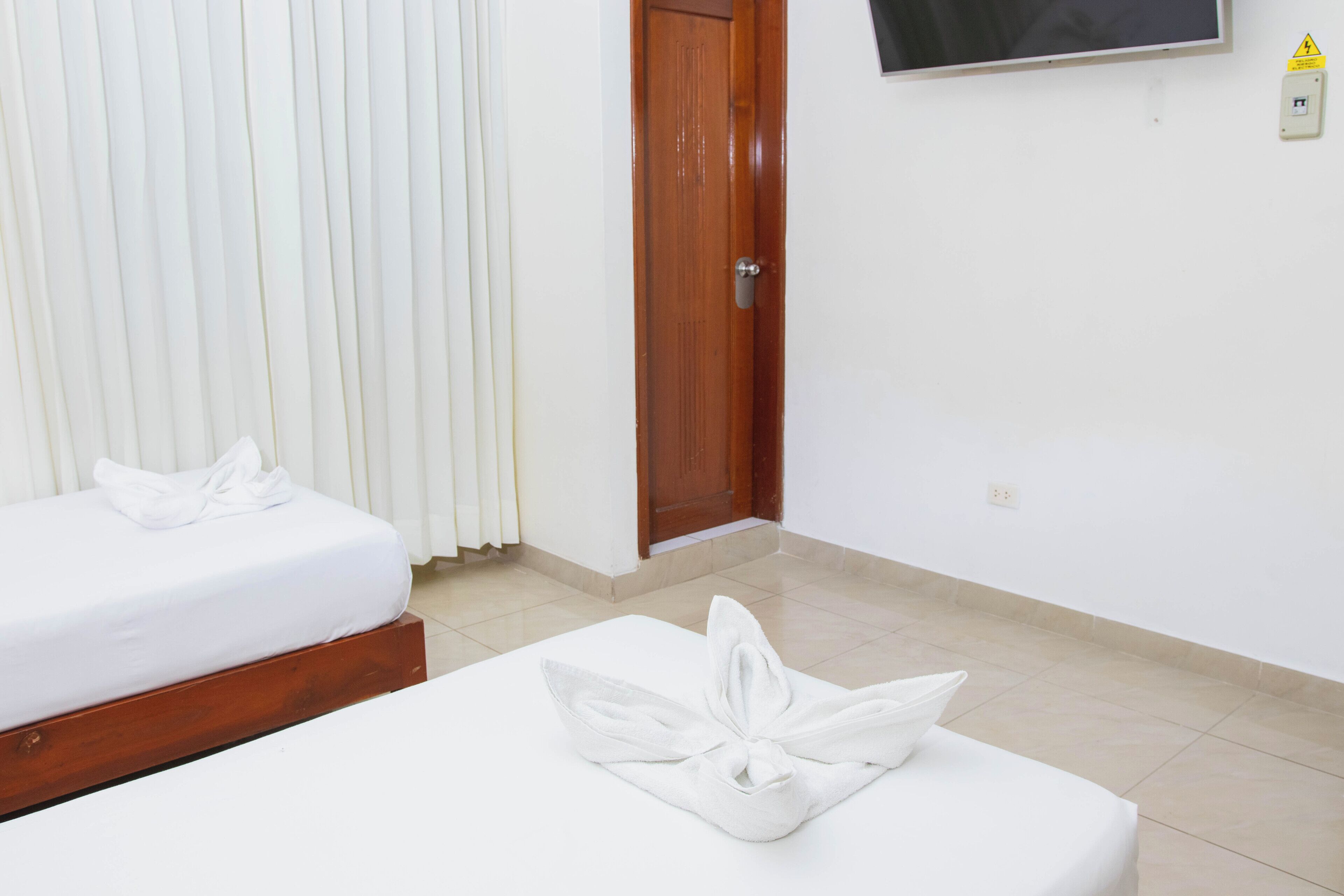 A hotel room with two beds, a wooden door, a white wall, and a television mounted above the door.